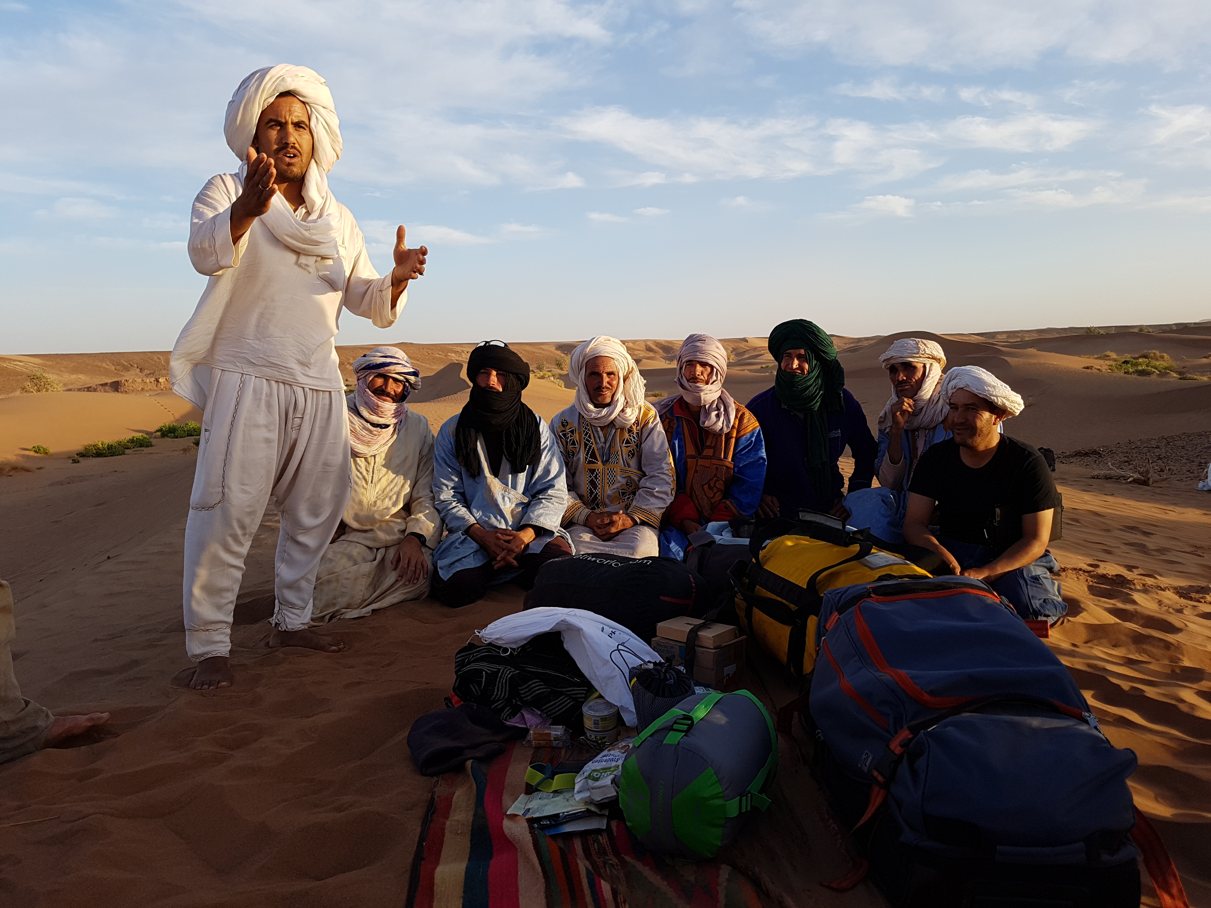 Nomaden sitzen auf einer Düne, vor ihnen steht Brahim und spricht in die Kamera